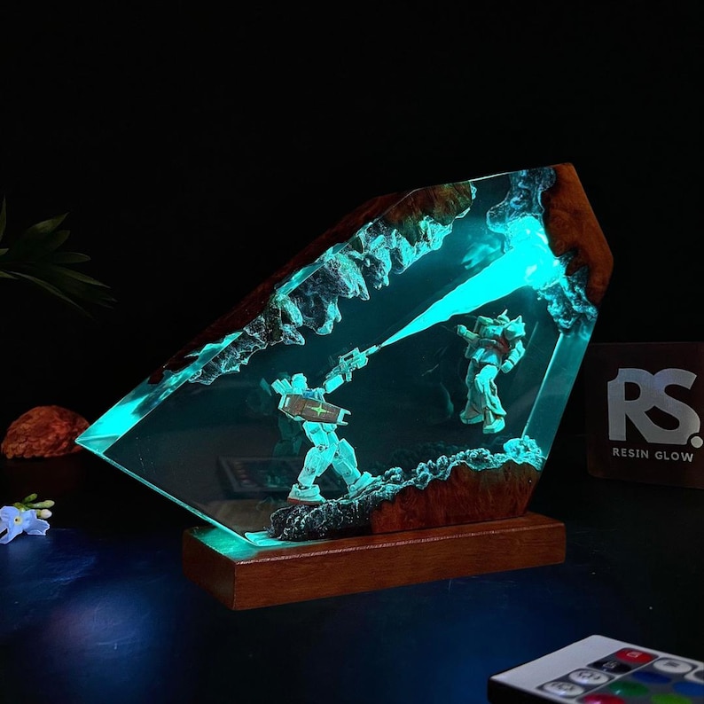 May include: A resin art piece featuring two robot figures in a battle scene, illuminated by a vibrant blue light. The artwork is encased in a clear resin block with a wooden base, creating a unique and eye-catching display.