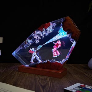 May include: A unique resin art piece featuring two robot figures in a battle scene, with a blue beam effect. The artwork is encased in a clear, angled resin block with a wooden base, and is illuminated with blue light.
