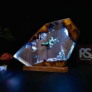 Puede incluir: Una pieza de arte decorativa de resina que representa a un mago en una escoba volando sobre un castillo. La obra de arte está encerrada en resina transparente con un marco y una base de madera. La pieza tiene un fondo estrellado y está iluminada con luz azul.