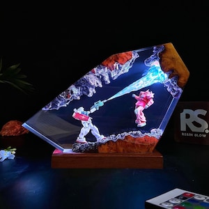 May include: A resin art piece featuring two robot figures in a battle scene. One robot is white, red, and blue, firing a blue energy beam at a pink robot. The artwork is set in a clear resin block with a wooden base. The text "RS RESIN GLOW" is visible.