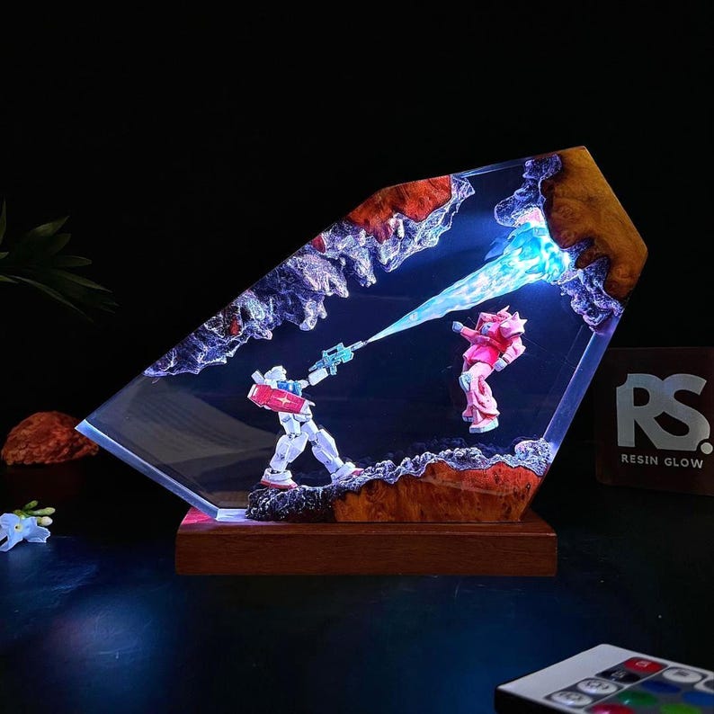 May include: A resin art piece featuring two robot figures in a battle scene. One robot is white, red, and blue, firing a blue energy beam at a pink robot. The artwork is set in a clear resin block with a wooden base. The text "RS RESIN GLOW" is visible.