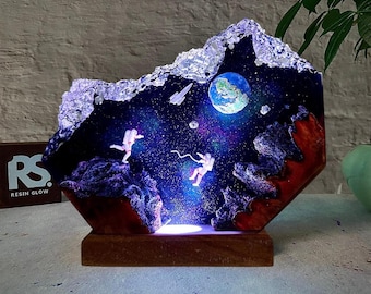 May include: A unique resin art piece depicting a space scene with astronauts, planets, and a galaxy. The artwork is illuminated with LED lights and set on a wooden base. The piece features a dark blue and purple color scheme with red and white accents.
