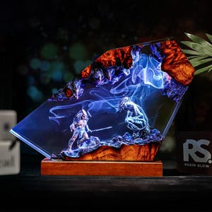 May include: A resin sculpture featuring a battle scene with two figures, set within a clear, angled block. The figures are detailed with a blue and gray color scheme, set against a backdrop of blue smoke and orange and brown accents. The sculpture is mounted on a wooden base.
