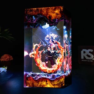 Könnte beinhalten: Ein rechteckiges Kunstwerk aus Kunstharz mit einer detaillierten Anime-Figur, umgeben von Flammen und anderen Charakteren. Das Kunstwerk ist in einem transparenten Kunstharzblock mit Leuchteffekt eingeschlossen, der lebendige Farben und komplizierte Details zeigt. Der Sockel hat eine holzähnliche Textur.