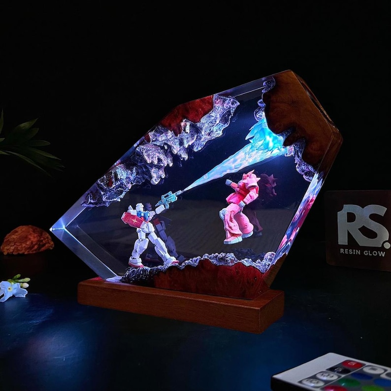 May include: A resin lamp featuring a battle scene with two robot figures, one in red, white, and blue, the other in pink. The lamp has a wooden base and a clear, angled resin body with embedded lights. The text "RESIN GLOW" is visible.