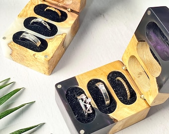 May include: Two wooden ring boxes with open lids, each holding three rings. The boxes have a natural wood grain with black and white resin accents. The rings are set in black velvet. One box has a gold ring, a diamond ring, and a silver ring.