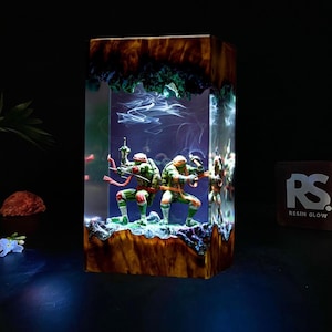 May include: A rectangular resin art piece featuring Teenage Mutant Ninja Turtles figures. The figures are in a clear resin block with a wood-grain base and a green and blue top. The piece is illuminated with a blue light.