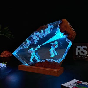 May include: A unique resin art piece featuring two robot figures engaged in battle, illuminated by blue light. The artwork is encased in a clear resin block with wooden edges and sits on a wooden base. The piece is a decorative item.