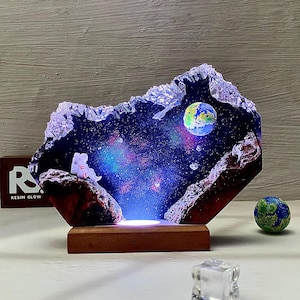 Könnte beinhalten: Eine Harz-Leuchte mit einer Galaxie-Szene, die eine kleine Astronautenfigur, einen Planeten und einen sternenklaren Nachthimmel zeigt. Die Lampe wird mit einem warmweißen Licht beleuchtet.