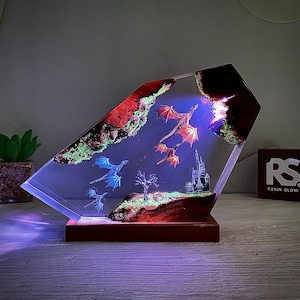 Puede incluir: Lámpara decorativa de resina con una escena de fantasía con dragones rojos y azules, un castillo y un árbol. La lámpara tiene una base de madera y un cuerpo transparente en ángulo. La escena está iluminada con luces azules y rojas.