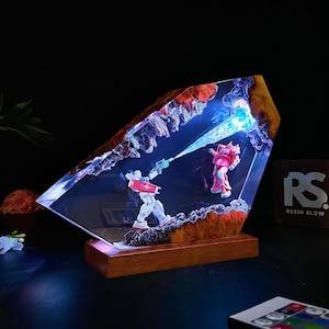 May include: A resin diorama featuring two robot figures in a battle scene, illuminated with blue light. The figures are set within a clear resin block with a wooden base and a textured, rocky border. The diorama has the text "Resin Glow".