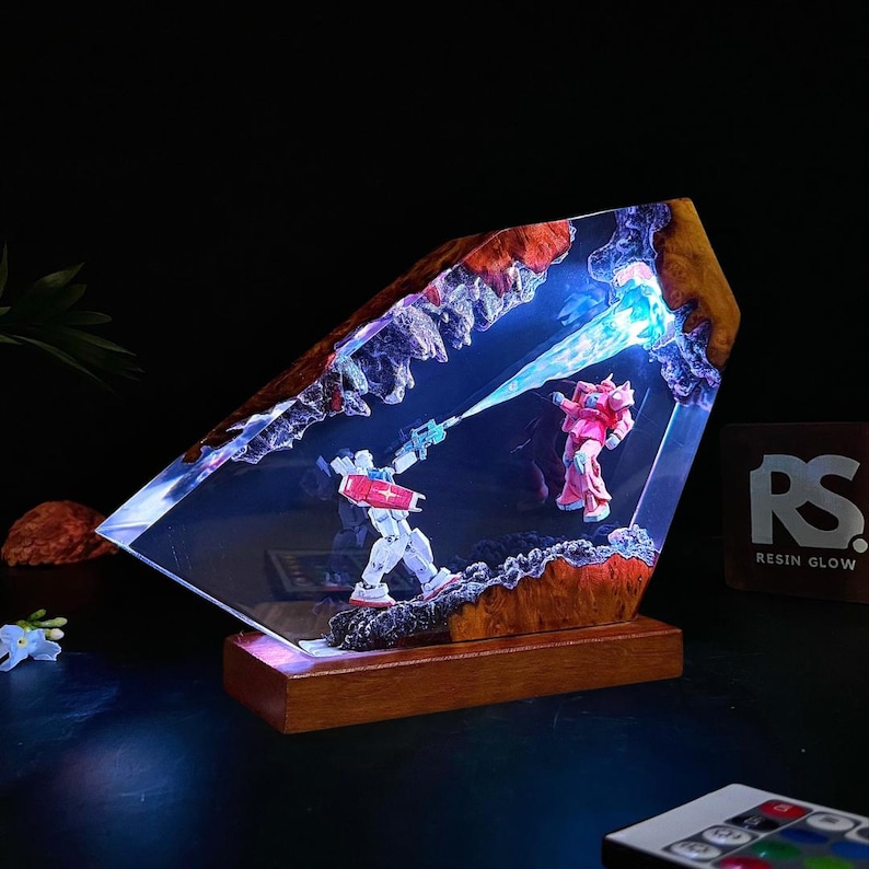 May include: A resin diorama featuring two robot figures in a battle scene, illuminated with blue light. The figures are set within a clear resin block with a wooden base and a textured, rocky border. The diorama has the text "Resin Glow".