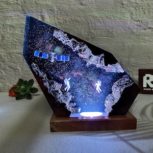 May include: A unique resin art piece depicting a space scene with astronauts and a satellite. The artwork is encased in a geometric shape, mounted on a wooden base, and illuminated with a soft, blue light. The colors include deep blues, purples, and white.
