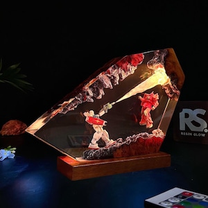 May include: A resin sculpture featuring two robot figures in a battle scene, illuminated by an internal light. The sculpture is set on a wooden base and has a jagged, rock-like border with red accents. The overall design is artistic and detailed.