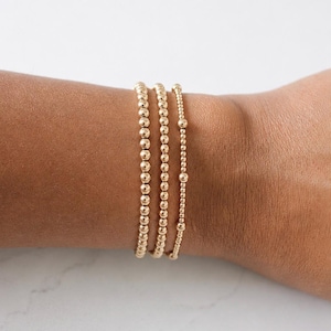 May include: A set of three gold-toned beaded bracelets worn on a wrist. Two bracelets feature uniform, medium-sized round beads, while the third bracelet displays a mix of smaller beads interspersed with three larger, distinct round beads.