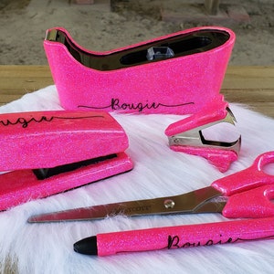 May include: A set of pink glitter office supplies, including a stapler, tape dispenser, staple remover, scissors, and pen. The word "Bougie" is written on the stapler, tape dispenser, and pen. The scissors have silver blades.