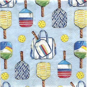 May include: Light blue napkin with a repeating pattern of pickleball paddles, balls, and a sports bag. The paddles feature various designs and colors, including red, white, and blue stripes, and yellow and blue patterns. Yellow pickleballs are scattered throughout.