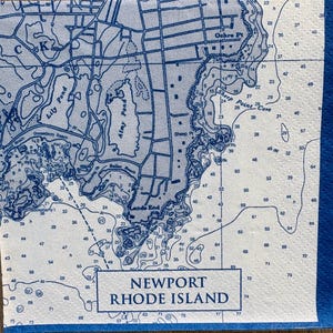 May include: A blue and white nautical map of Newport, Rhode Island, featuring detailed coastlines, landmarks, and depth soundings. The map includes the text "Newport Rhode Island" at the bottom, and is likely printed on a napkin.