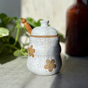 May include: A speckled white ceramic sugar bowl with a lid and a wooden spoon. The bowl features two brown flower designs. The spoon has a wooden handle. The bowl is approximately 4 inches tall.