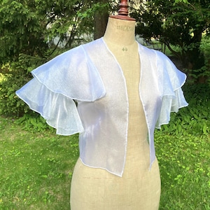 May include: A sheer, light blue bolero jacket with ruffled sleeves. The open-front design is displayed on a mannequin. The fabric has a subtle sheen, and the garment is lightweight, suitable for layering. The background features green foliage.