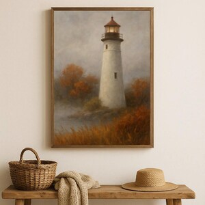 Puede incluir: Impresión enmarcada de un faro blanco con tejado rojo, rodeado de follaje otoñal. Una cesta, una manta y un sombrero están sobre un banco de madera en primer plano. Pintura en tonos apagados.