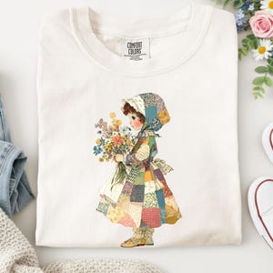 Op de afbeelding: Crèmekleurig T-shirt met een vintage illustratie van een meisje in een patchwork jurk met een boeket bloemen. Het shirt heeft een Comfort Colors label. Witte sneakers en een spijkerbroek zijn zichtbaar.