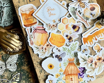 May include: A set of bee-themed stickers featuring honeycombs, bees, jars of honey, flowers, and a beehive. The stickers are on a vintage book with the text "Bee Happy" and "Bee You".