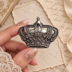 May include: A silver-toned metal crown brooch featuring an antiqued finish with intricate embossed floral details, a central finial, and a band decorated with a repeating diamond pattern, showcasing a classic regal design with visible metallic texture.