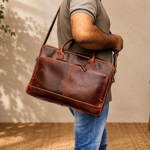 Puede incluir: Un maletín de cuero marrón con una correa para el hombro y un asa superior. El maletín tiene un bolsillo frontal y costuras visibles. La bolsa es llevada por una persona que lleva una camisa gris y vaqueros azules.