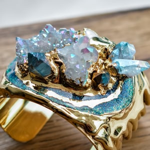 May include: A gold-colored cuff bracelet with a large, ornate design. The bracelet features a central cluster of iridescent crystals in shades of blue, white, and purple. The cuff has a textured, organic shape with a metallic finish.