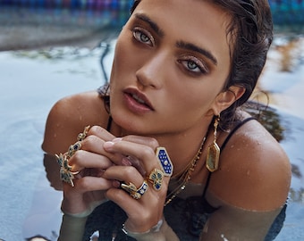 May include: A woman wearing a variety of gold rings with blue gemstones, including a large ring with a geometric design. She is also wearing a gold necklace and earrings with blue gemstones.