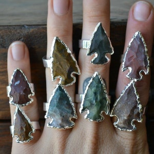 May include: A set of eight silver rings featuring arrowhead-shaped stones in various colors and patterns. The stones are set in silver and have a polished finish. The rings are worn on a person's fingers.