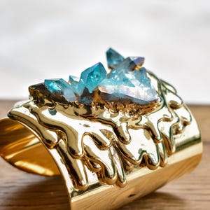May include: A gold-colored cuff bracelet with a textured, molten metal design. The bracelet is adorned with a cluster of blue-green crystals. The jewelry is displayed on a wooden surface.