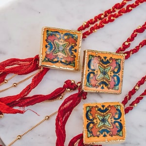 May include: Three gold-framed square pendants with colorful tile-like patterns on a red and gold chain. The pendants feature intricate designs with blue, green, red, and yellow colors.