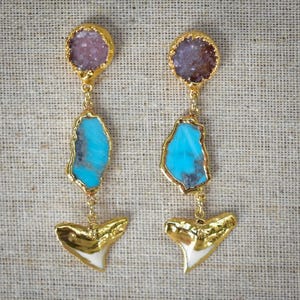 Pode incluir: Um par de brincos dourados com uma parte superior de druzy, uma pedra turquesa e um design de dente de tubarão. Os brincos são suspensos por uma corrente dourada. O fundo é de cor bege neutra.