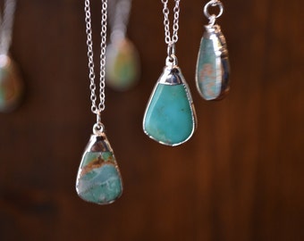 May include: Close-up of several silver necklaces with teardrop-shaped pendants. The pendants feature turquoise-coloured stones with silver trim. The necklaces hang against a blurred brown background, showcasing the jewellery's design and colour.