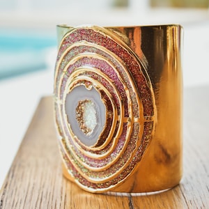 May include: A gold-colored cuff bracelet with a large, circular design. The design features concentric rings of glitter in shades of pink, red, and gold, surrounding a central, light-colored stone. The bracelet is displayed on a wooden surface.