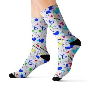 Peut inclure: Une paire de chaussettes montantes blanches avec un motif coloré composé de cœurs bleus, jaunes et roses, de rubans et du texte T21 dispersés. Les chaussettes présentent des talons et des pointes noirs unis, contrastant avec le tissu à motifs.