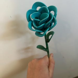 Puede incluir: Una rosa de metal azul turquesa con tallo y hojas de metal verde. La rosa está hecha de múltiples capas de metal que están curvadas y superpuestas.