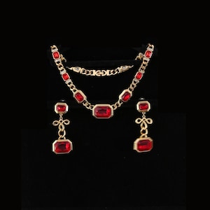 Puede incluir: Un conjunto de joyas doradas que incluye un collar, una pulsera y pendientes. El collar y la pulsera están adornados con piedras preciosas rectangulares de color rojo rubí. Los pendientes tienen un diseño similar, con piedras preciosas rojo rubí más pequeñas.
