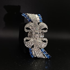 Può includere: Spilla argentata con design a fiocco, incastonata con cristalli trasparenti e blu. La spilla presenta un gruppo rettangolare centrale e dettagli di cristalli blu a cascata, su sfondo scuro.