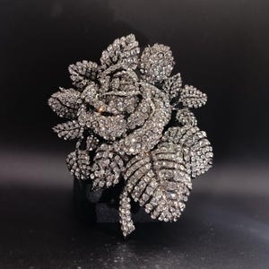 Puede incluir: Broche plateado con forma de rosa, incrustado con numerosos cristales brillantes. El broche presenta hojas y pétalos detallados, creando un diseño floral realista. Se encuentra sobre un fondo oscuro, resaltando su brillo y detalles.