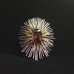 May include: An eye-catching brooch with a central, rectangular, deep red gemstone. The brooch features a sunburst design with alternating gold and silver-toned metal spikes radiating outwards from the gemstone, set against a black background.
