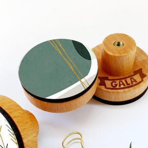 Könnte beinhalten: Drei Holzknöpfe mit verschiedenen Designs. Der erste Knopf hat ein abstraktes Design in Grün und Weiß mit goldenen Akzenten. Der zweite Knopf hat einen weißen Hintergrund mit einem Muster aus goldenen und grünen Pfeilen. Der dritte Knopf hat das Wort "GALA" eingraviert.