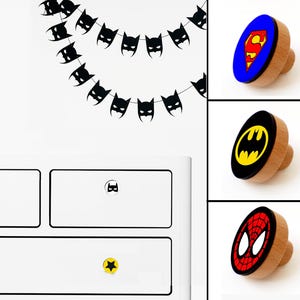 May include: Three wooden drawer pulls featuring superhero logos. The first pull has a red and yellow Superman logo on a blue background. The second pull has a black and yellow Batman logo on a black background. The third pull has a red and black Spiderman logo on a red background.