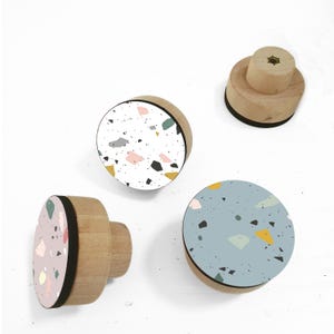 May include: Three round wooden knobs with a terrazzo pattern. The knobs are in pink, blue, and white with black, yellow, and green accents.
