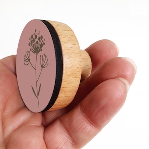 May include: A wooden stamp featuring a floral design. It has a light brown wooden handle and a circular stamp face. The stamp face is a dusty rose colour with a brown floral illustration. A black band separates the stamp face from the wooden handle.