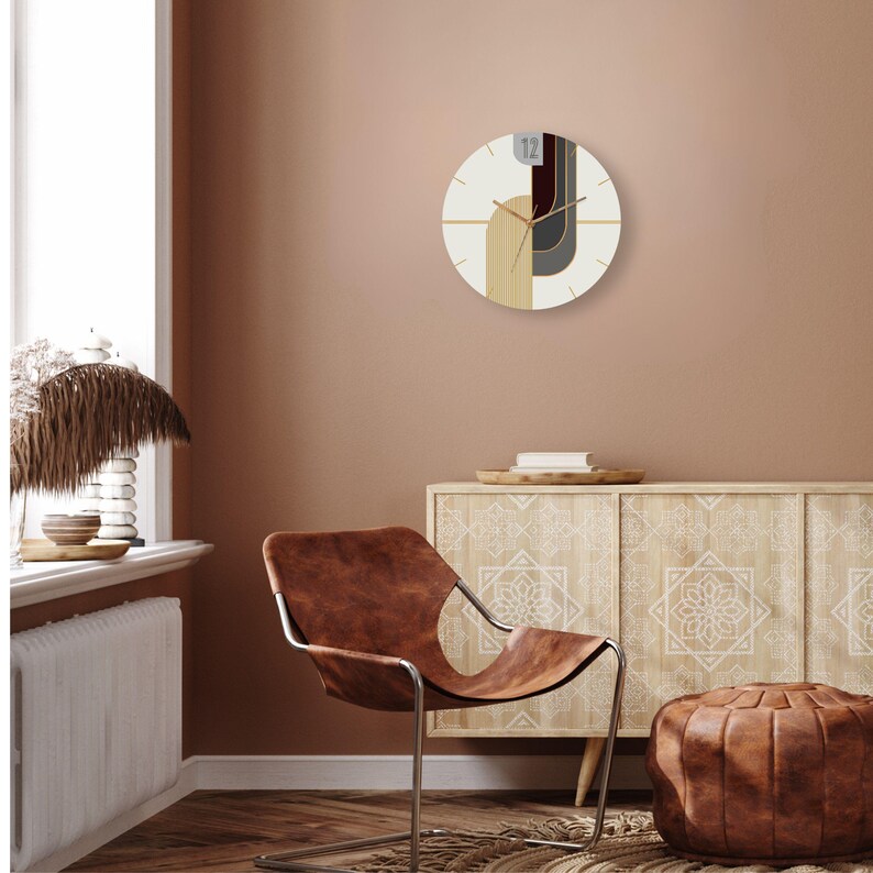 May include: A white and brown geometric wall clock with black and gray accents. The clock is hanging on a light brown wall above a wooden cabinet with a white floral design. A brown leather chair and ottoman are in the foreground.