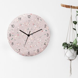 May include: A pink round clock with black hands and black numbers on a white wall. The clock face has a terrazzo pattern with pink, white, and grey flecks.