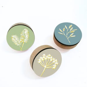 Puede incluir: Tres pomos de armario de madera con diseños botánicos. Uno es verde claro con hojas blancas y amarillas en forma de helecho. Otro es gris con una flor blanca y amarilla. El tercero es azul con un diseño de hojas blancas y amarillas.